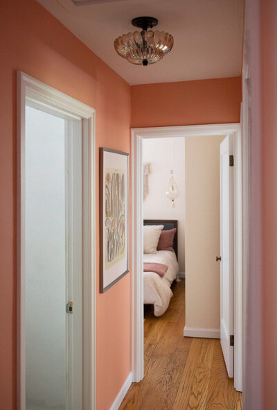 A hallway painted in pink with artwork hanging on the wall and a peek into a bedroom.