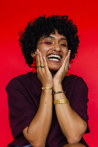 A color portrait of a woman with a big smile on her face with both hands touching her cheeks. The backdrop is a deep red