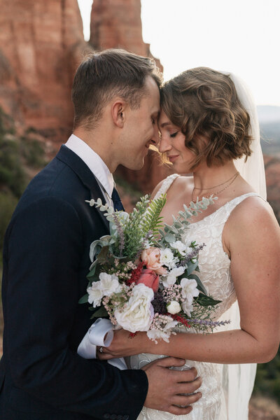 Close-up portrait of a bride and groom embracing during their elopement at Cathedral Rock in Sedona, Arizona, with red rock formations and a romantic bouquet in view.