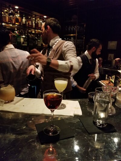 A perfectly crafted Manhattan cocktail served in a Nick & Nora glass with three cherries, set on a dimly lit bar with expert bartenders in the background.