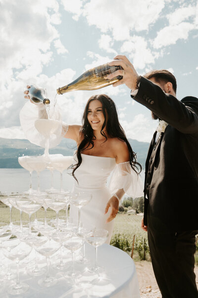 KChampagne Tower Photo on Wedding Day in Kelowna BC, pouring the champagne from their venue vineyard winery. elowna Wedding Couple over looking the Okanagan Lake in Lake Country BC.  They are wearing modern sleek wedding attire with a gorgeous wedding dress and black tux.  A huge floral arch behind them filled with babysbreath.  A modern wedding day with a gorgeous look out!  They are dancing in their reception at the 50th Parallel Estate