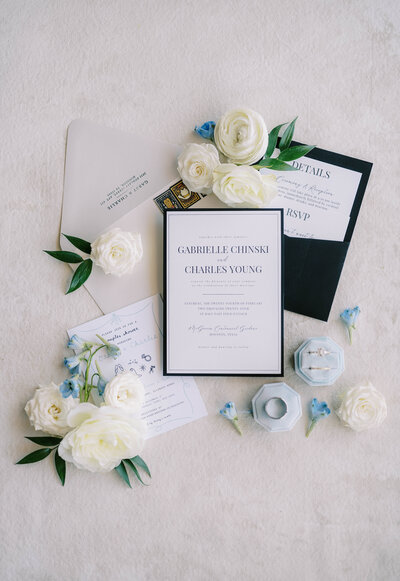 Flat lay of white and black wedding invitation suite with rings and blue and white flowers