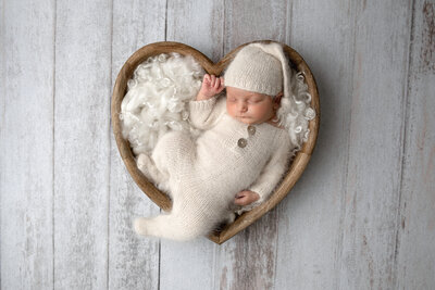 Newborn baby peacefully sleeping in a wooden heart-shaped bowl, dressed in a soft cream knit outfit with a matching hat, surrounded by white wool accents. A serene and timeless setup from a professional newborn photography session