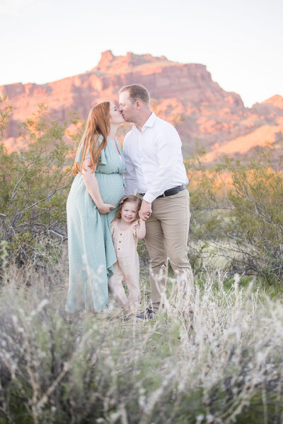 Pregnant wife and husband kissing while holding toddler daughter's hand in a desert landscape