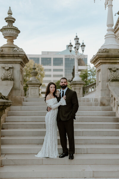 A couple poses on elegant stone steps flanked by ornate lampposts. The woman wears a white lace dress, and the man is in a dark suit, exuding a formal and romantic tone.