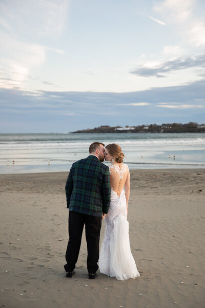 Rhode Island Wedding Photographer | Kelsey Sheehan Photography Timeless Rhode Island Weddings | A couple stands on a sandy beach at sunset, the woman in a white gown and the man in a dark suit. They lean in close, gazing at the serene ocean.