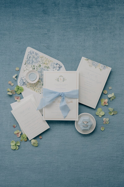 Wedding invitation with a blue background and hydrangeas surrounding, with blue ribbon. Photo by Anna Brace who specializes in Nebraska Wedding Photography
