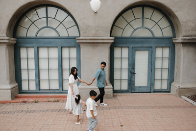 A stylish family of four walks hand in hand in front of arched blue-framed windows during a modern family photo session.