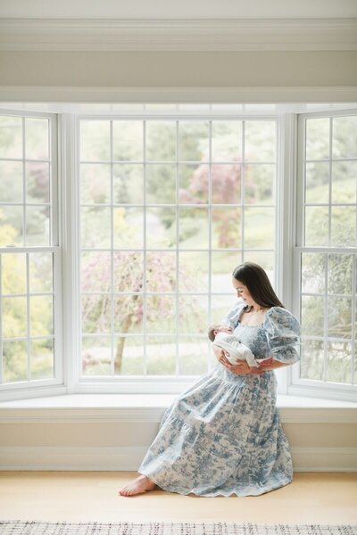 a mother in a blue floral dress sits by a window holding her newborn baby during NJ newborn photos at home.