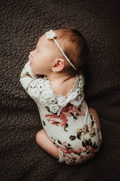 A newborn girl wears a floral romper lying on a gray blanket.