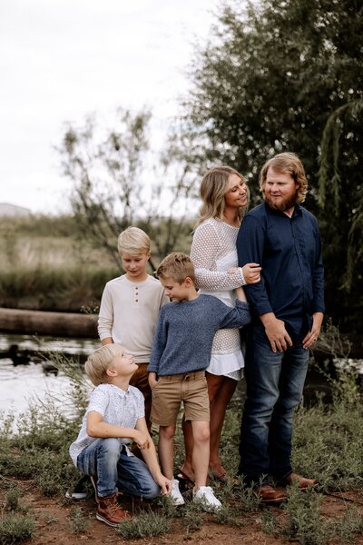 family of five standing in front of a family pond. Family colors for this session are navy and white. Mom is hugging onto dad and the boys are smiling looking at each other. Captured by Luboock family photographer Mary Wiebe