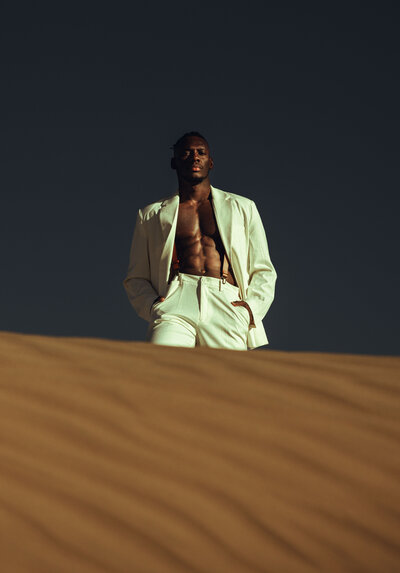 Man standing on sand, dark blue skies, brown sand, white pant suit, posing,