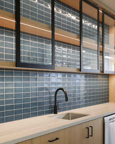 A kitchen counter and sink area featuring a blue tile backsplash, wood cabinetry with black accents, and open shelving with movable glass panels