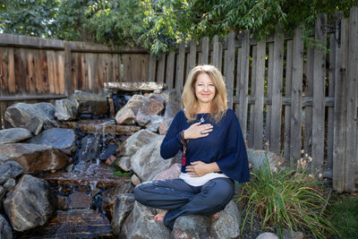 Cyndi sitting in her garden in lotus position, Her right hand positioned on her heart and her left hand positioned on her belly.