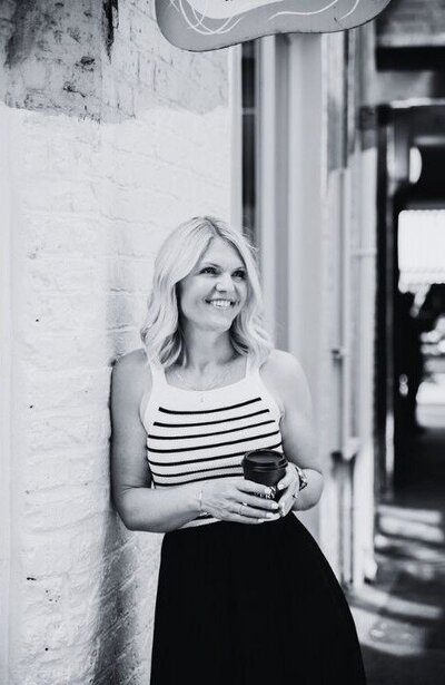 Emily Collins smiling and holding a coffee cup while leaning against a brick wall in a black and white photo