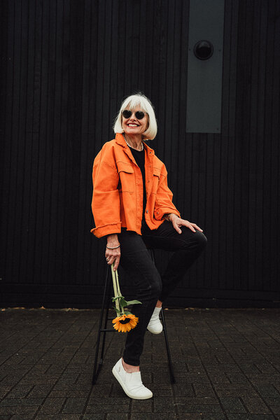 Personal brand image of woman wearing bright orange jacket and holding sunflower in front of contrasting black background