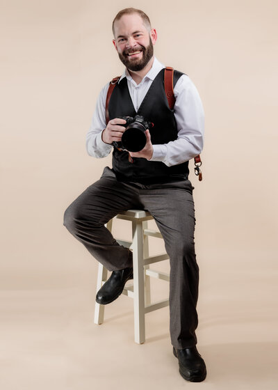 White Male Lehigh Valley  Wedding photographer Eric Boylan wearing a holdfast harness and black vest  holding a Canon R6 while sitting on a stool against a beige backdrop