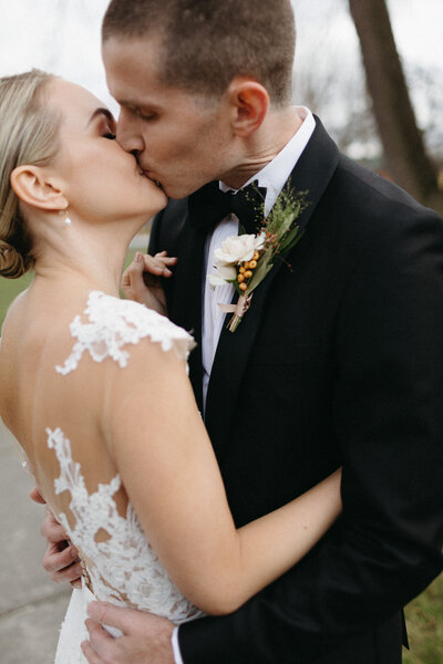 A bride and groom share a kiss outdoors, captured beautifully by Jennifer van Son, a wedding photographer in Toronto at Cape Picton. The bride wears a lace gown, and the groom is in a dark suit with a boutonniere.