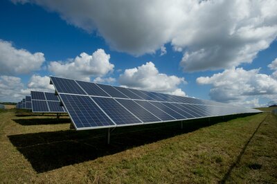 Wide-angle view of a solar farm with multiple photovoltaic panels on a grassy field, generating renewable energy under a bright, partly cloudy sky