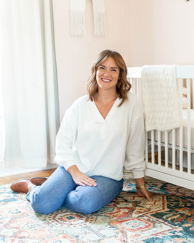 In-home location consultant, Sarah, sitting on the floor in a nursery next to a crib wearing a white blouse and jeans