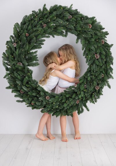Image of two sisters kissing on a Christmas wreath backdrop in the studio. Taken by Norwich portrait photographer Claire Howes. Christmas Mini Sessions Norwich.