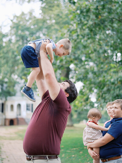 A dad lifting up a toddler with other kids watching them during a family photography session in Richmond by Katie Stansfield Photography.