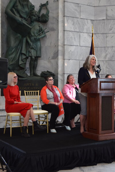 Celeste Mergens speaking at a State Capital event. Woman standing at podium speaking while others look on.