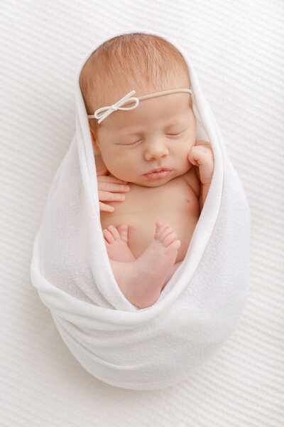 Sleeping Newborn baby  wrapped in a white swaddle with feed and hands and chest showing. She has a little minimalist white bow on her head and she is laying on a white textured blanket.