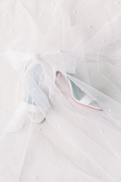 A bride's white wedding shoes underneath a sheer white veil with pearls