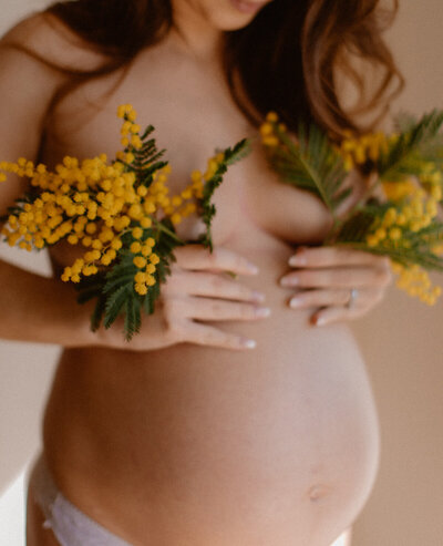 Une femme enceinte en culotte qui couvre sa poitrine par du mimosa dans chacune de ses mains.