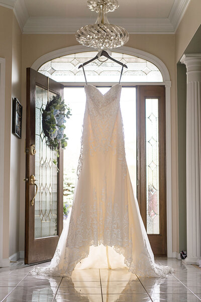 Wedding dress hangs from wooden hanger in bright and airy foyer filled with natural light