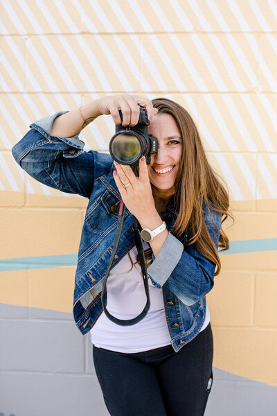 denver branding photos of a woman standing holding a nikon camera against a yellow mural wall wearing a jean jack and black pants