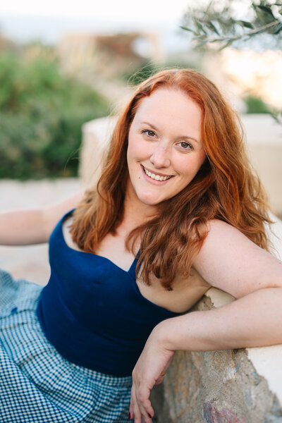 Reheaded woman leans on a ledge while seated and smiles into the camera