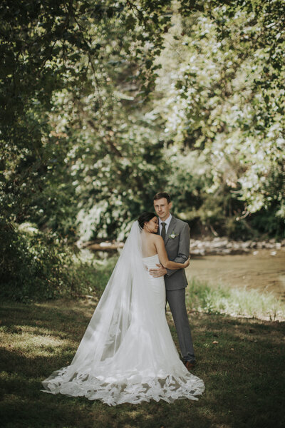 A bride and groom embrace outdoors beside a quiet stream, surrounded by lush green trees and soft sunlight filtering through the leaves.