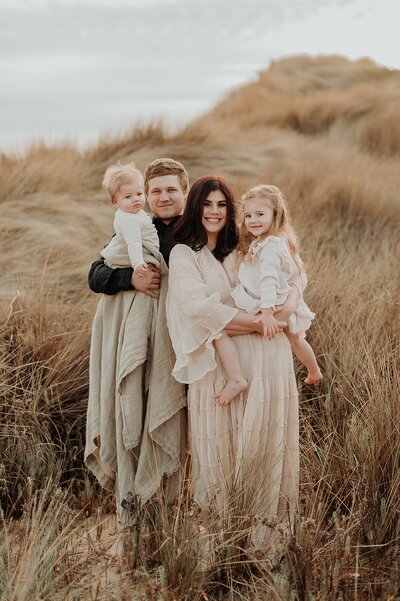 Family of 4 poses in a field for a family photo session. Each parent is holding a young child.  Everyone is dressed coordinating outfits in neutral soft ones.