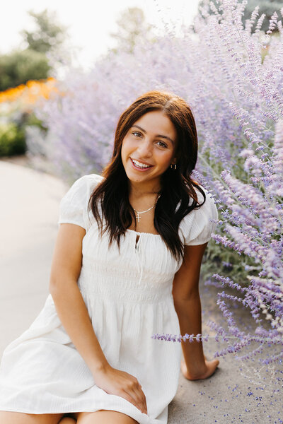 Senior girl in a white dress sitting on the ground in front of purple floral bushes