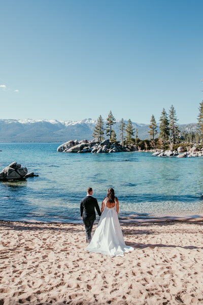 Married couple walking barefoot along a sandy beach at Lake Tahoe, smiling at each other with tall pine trees and calm water in the background.