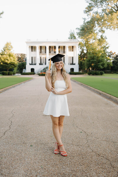Graduate in white dress posed with graduation cap in front of the President's Mansion at the University of Alabama