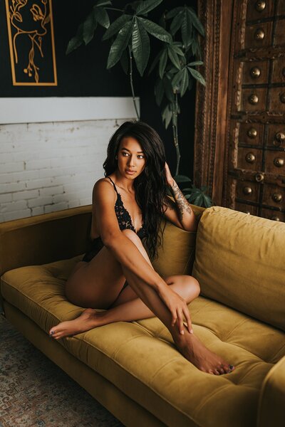 Denver boudoir photography of a black woman sitting on a yellow sofa wearing a black one-piece, with her hand in her hair