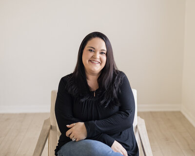 Woman sitting in a chair with legs crossed, has black hair, wearing a black shirt, blue jeans, and black shoes, smiling at the camera