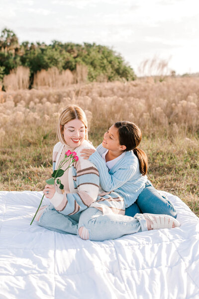 Mom sits on a bed in the middle of a beautiful pampas field with her daughter during their phtoography session in an Orlando field.