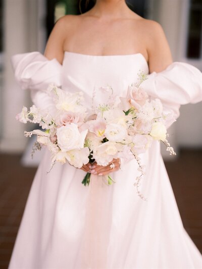 A bride in an off-shoulder white gown holds a bouquet of light pink and white flowers, curated beautifully by a top Canadian wedding planner.