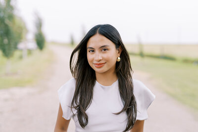 A wedding designer with long dark hair stands, wearing a white top stands outside on a dirt road, looking directly at the camera.