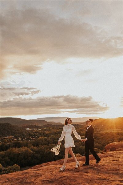 A couple walks hand in hand across a rocky ledge at sunset during their elopement in Sedona, Arizona. One partner is wearing a short white dress, and the other is in a black suit, with a stunning view of the landscape and a golden sky behind them.