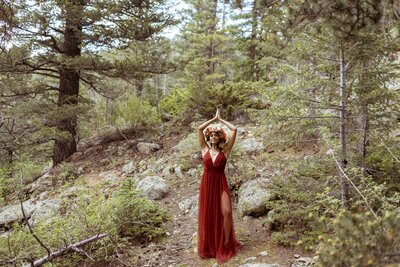 woman with a flower crown and a red dress standing in the forest with her hands joined above her head