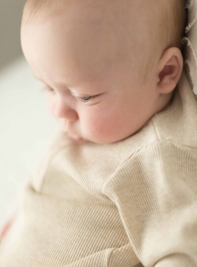 Close-up of a baby in a soft beige outfit, gazing downward in a quiet moment. A timeless baby session by Moments in Light Photography.