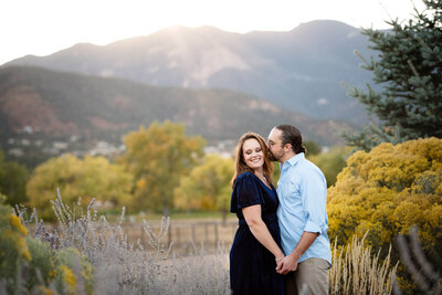 Guy kissing his fiancé on the cheek outside by fall foliage with a Colorado mountain backdrop