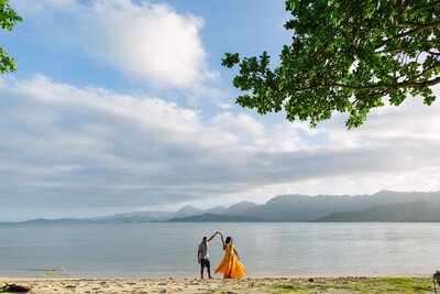 A man and a woman wearing a yellow dress dance on the beach.