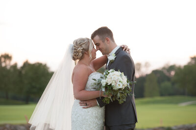 Bride holding white bouquet touching foreheads with groom in a light gray tuxedo with the Washington Missouri river view in the background at Riverbend Chapel