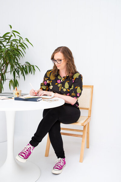 Melissa-Packham_A-Brand-Is-Not-A-Logo_Melissa-working-while-sitting-at-table-on-cane-chair-plants-in-corner_wearing-floral-top-black-jeans-magenta-converse-sneakers_Portrait